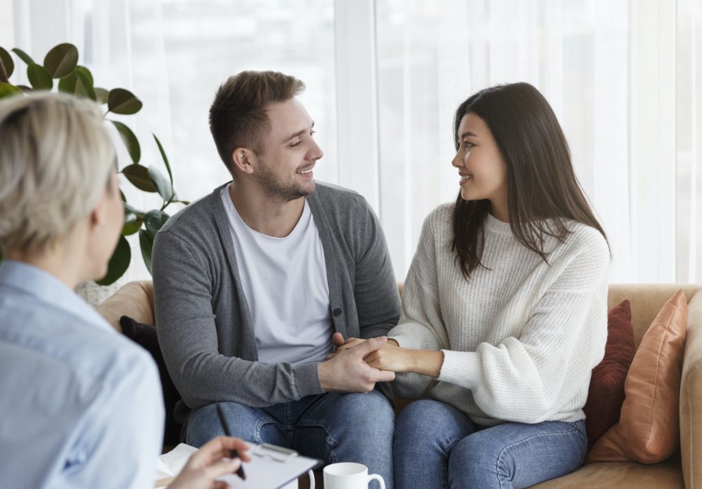 Happy Couple Holding Hands Reconciling After Therapy Sitting In Office